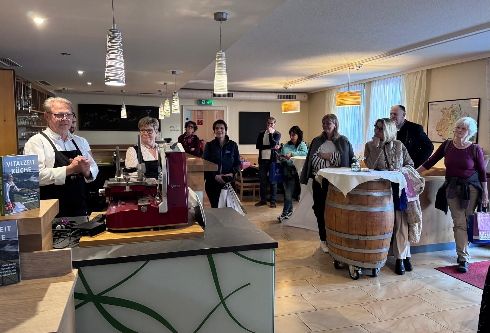 Rund 8 Personen stehen in einer Gaststube und hören den Gastgebern, die hinter der Bar etwas erzählen, zu. 