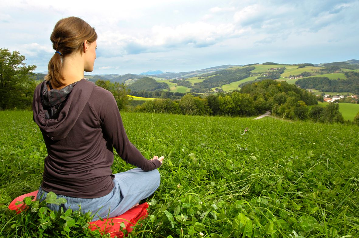 Eine Frau sitzt meditierend auf einer Wiese mit Blick auf eine hügelige Landschaft.