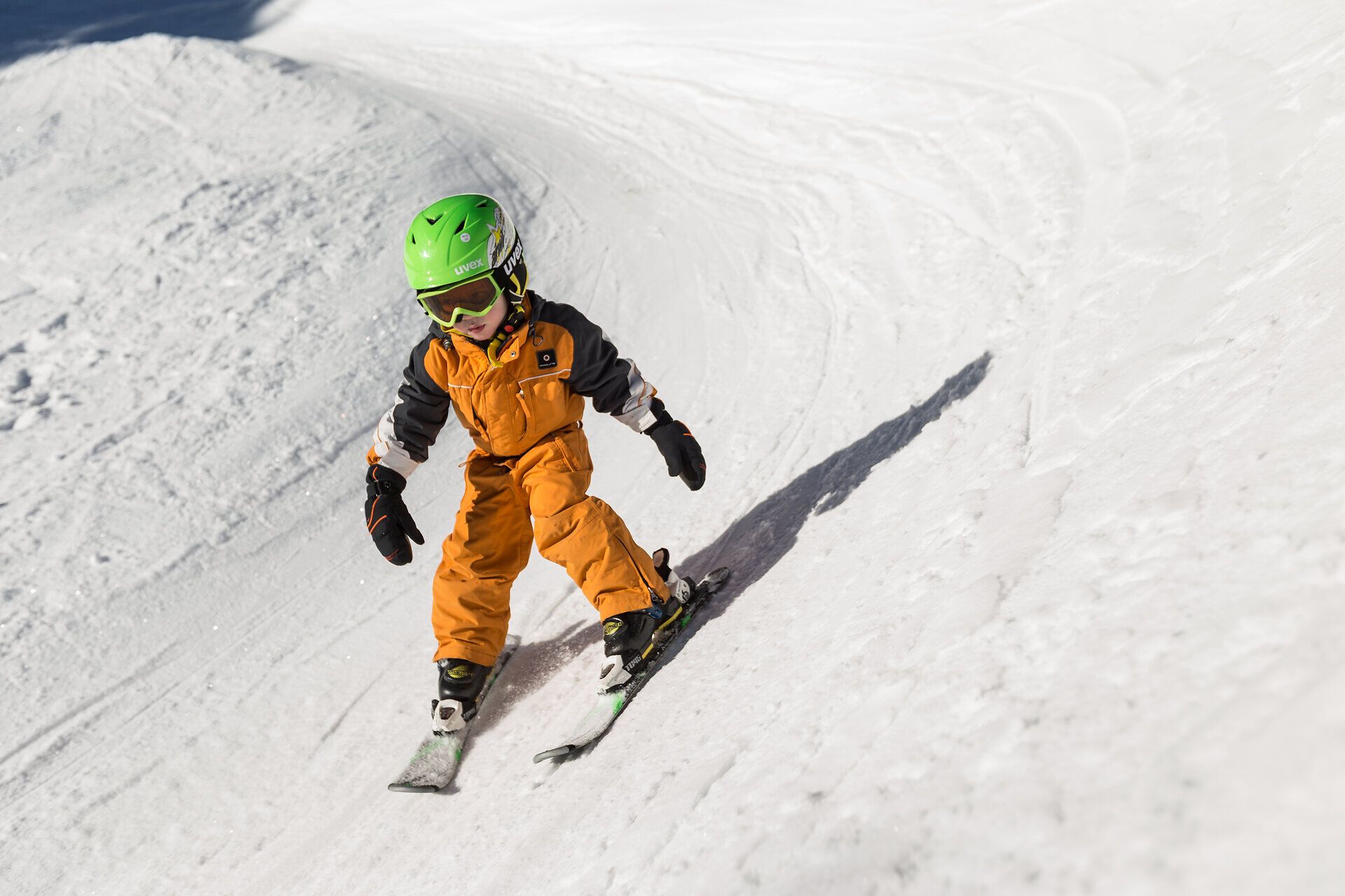 Ein kleiner Skifahrer gleitet mit Freude über die schneebedeckten Pisten, während die strahlende Sonne den Wintertag erhellt. In seiner leuchtend orangefarbenen Skijacke und dem grünen Helm strahlt er vor Begeisterung und Abenteuerlust. Die frische Bergluft und die schneebedeckte Landschaft schaffen eine unvergessliche Atmosphäre für Familien und Kinder.
