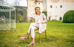 Frau sitzt auf einer Wiese vor einem historischen Schlossgebäude und blickt lächelnd in die Kamera.