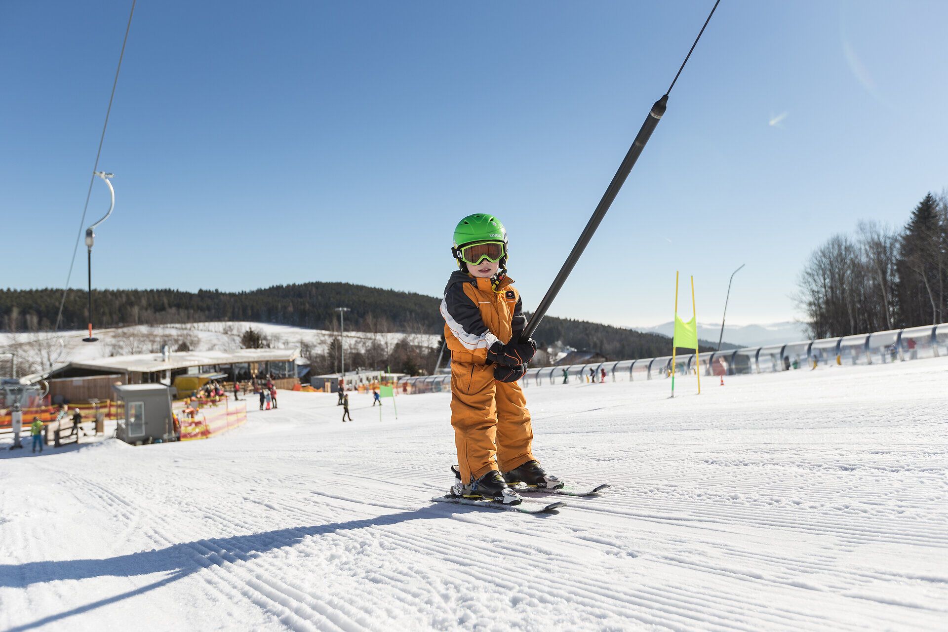 Ein strahlend blauer Himmel umrahmt die fröhlichen Szenen auf der Piste, wo Kinder mit Begeisterung ihre ersten Schwünge im Schnee meistern. Die frische Bergluft und die strahlende Sonne schaffen eine einladende Atmosphäre für Familien, die gemeinsam unvergessliche Momente erleben.