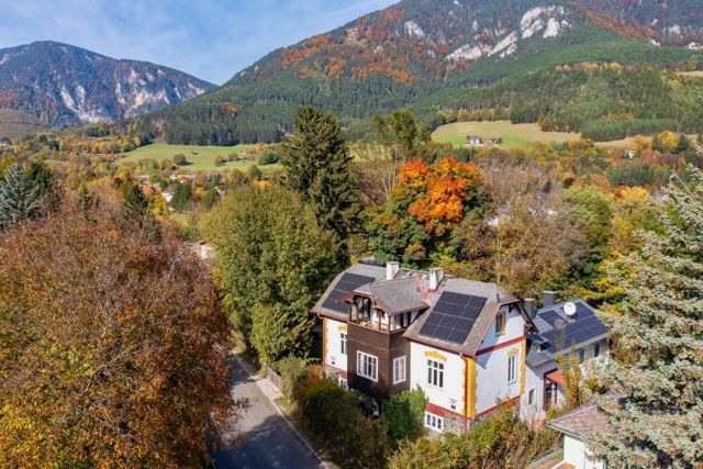 Luftaufnahme eines Landhauses mit Solarpanelen auf dem Dach, umgeben von herbstlichen Bäumen und Bergen im Hintergrund.