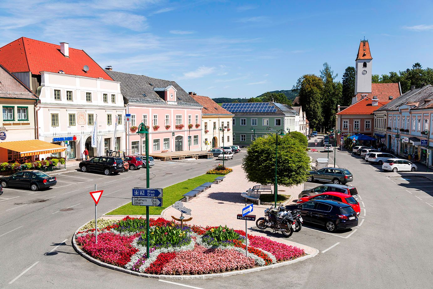 Hauptplatz in Aspang Markt mit bunten Blumenbeeten, geparkten Autos und historischen Gebäuden.