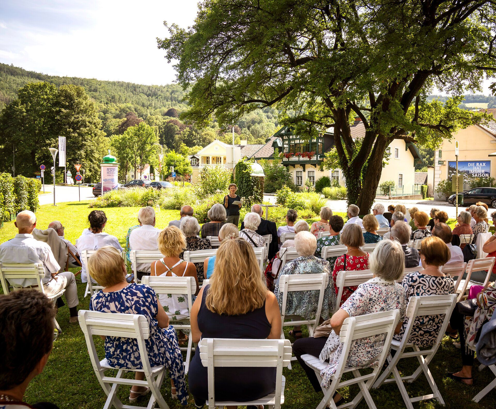 Unter dem schattenspendenden Baum versammeln sich zahlreiche Besucher, um den Klängen der Festspiele zu lauschen. Die sanfte Brise trägt den Duft von frischen Blumen und der grünen Wiesen in die Luft, während die Sonne die Szenerie in warmes Licht taucht.