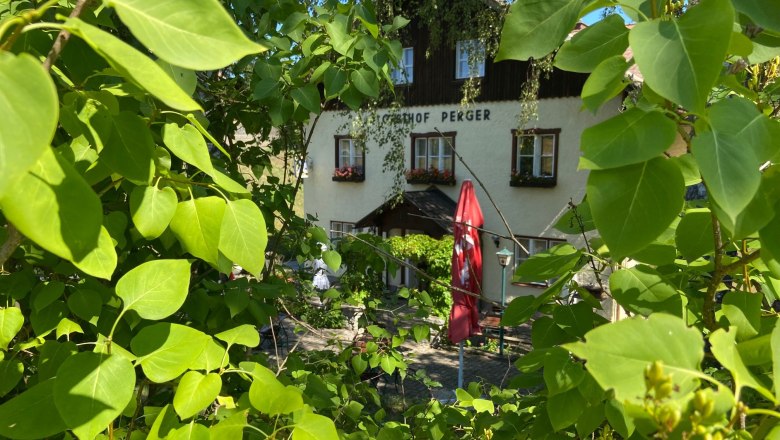 An inn with the inscription 'Gasthof Perger' behind green leaves and a red parasol.