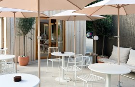 Moderne Terrasse mit weißen Tischen und Stühlen, Sonnenschirmen und Holzhütte im Hintergrund.