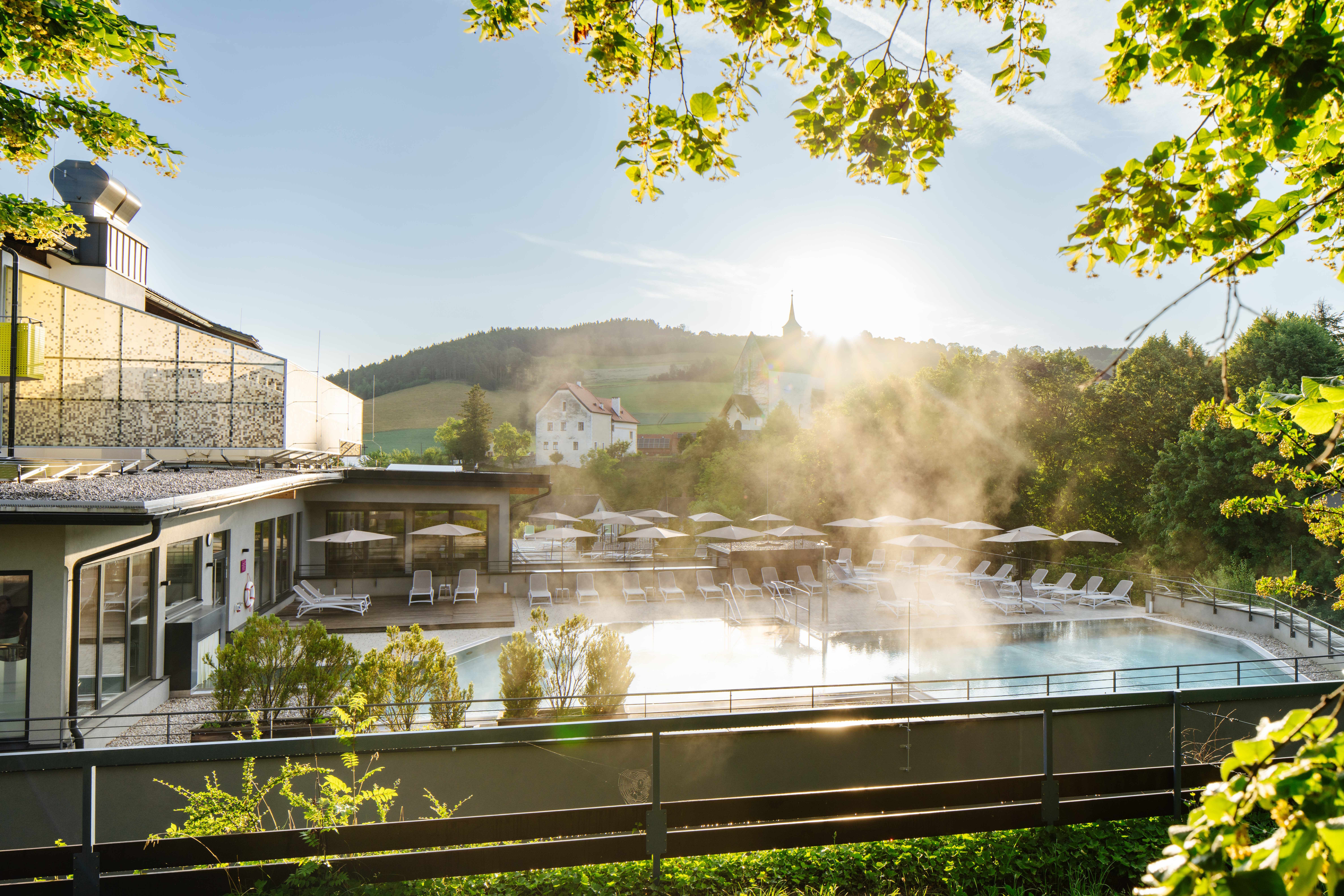 Ein Außenpool mit Liegestühlen und Sonnenschirmen, umgeben von grüner Landschaft und einem Gebäude im Hintergrund bei Sonnenaufgang.