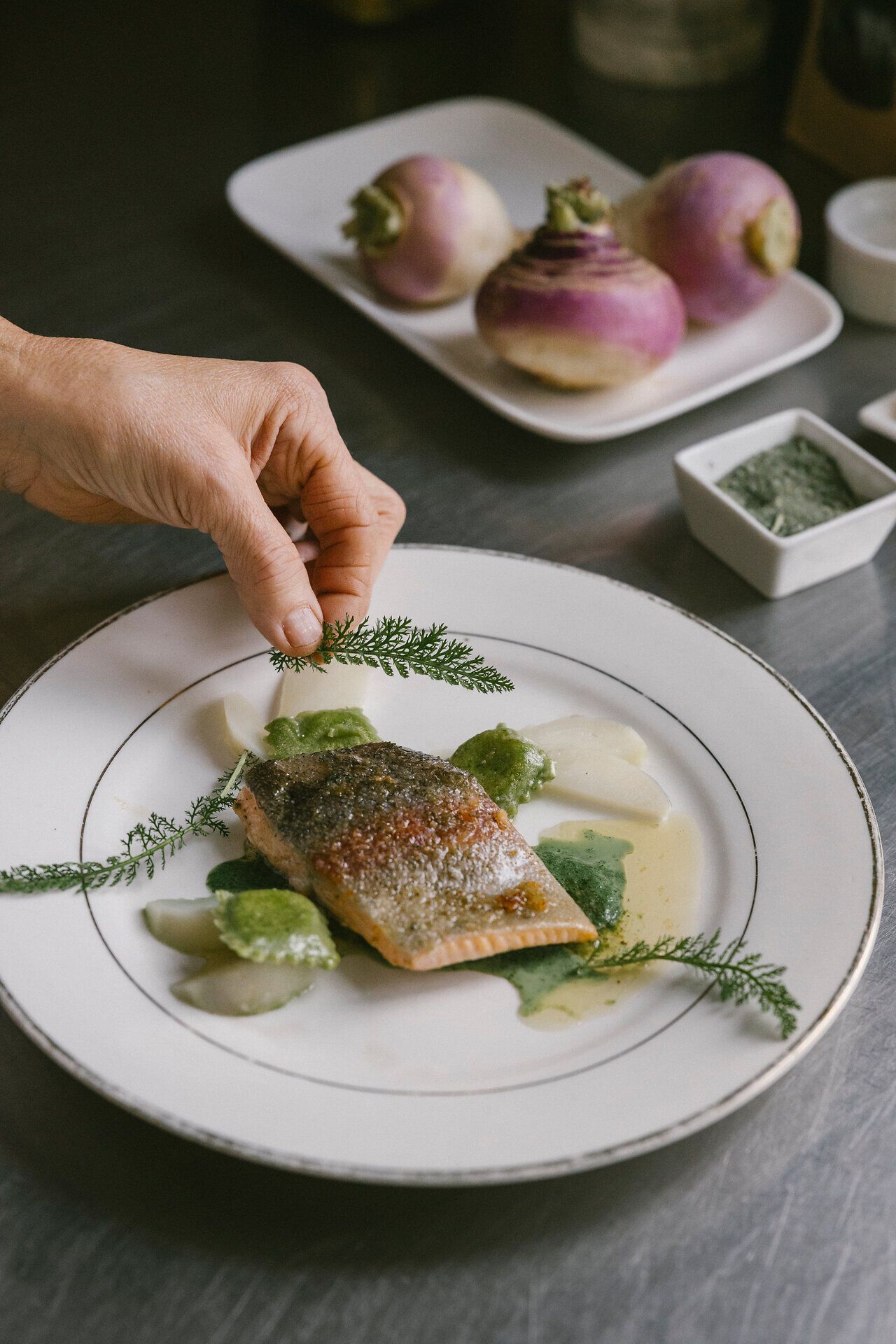Angerichtetes Fischfilet mit grünen Ravioli, hellen Gemüsestücken und frischen Kräutern auf einem weißen Teller.