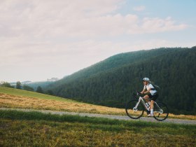 Eine Radfahrerin f&auml;hrt auf einem schmalen, steigenden Weg, im Hintergrund bewaldete H&uuml;gel.