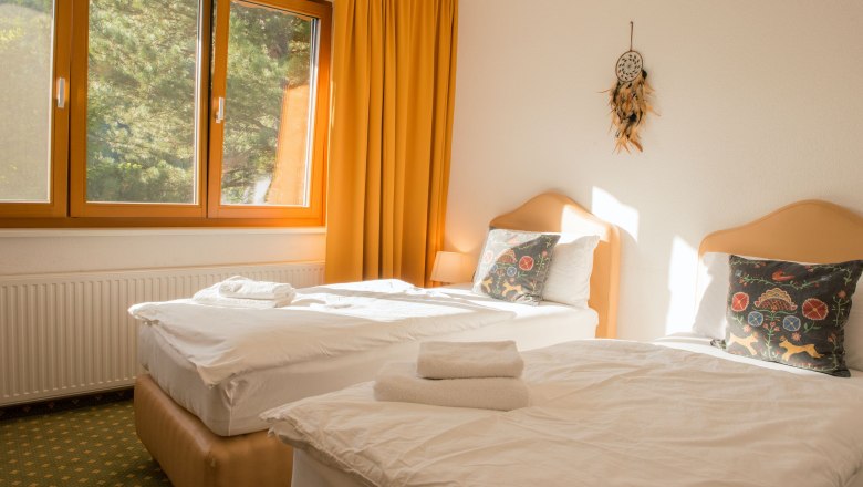 Ein helles Doppelzimmer mit zwei Einzelbetten, gelben Vorhängen und einem Fenster mit Blick ins Grüne.