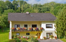 Zweistöckiges gelbes Haus mit Balkon und Blumen, umgeben von Bäumen und Garten.