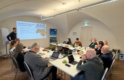 Mehrere Workshop Teilnehmer sitzen an einem Tisch während ein Mann einen Vortrag hält.