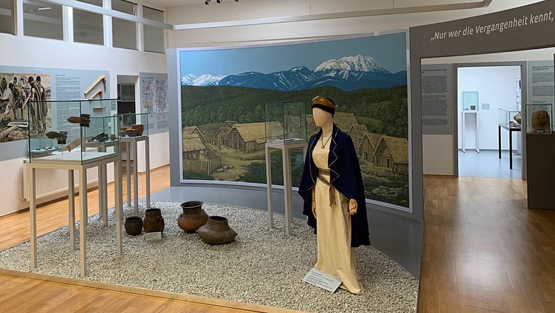 Ausstellung im Pittener Regionsmuseum mit historischen Artefakten und einer Figur in traditioneller Kleidung vor einem Wandbild.