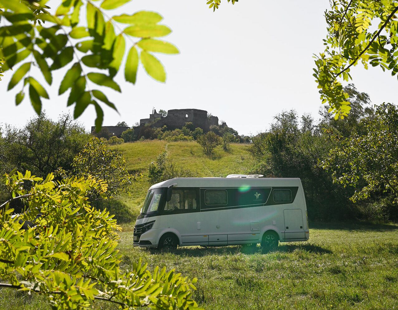 Umgeben von üppigem Grün und unter einem strahlend blauen Himmel, bietet der Campingwagen einen perfekten Rückzugsort für Naturliebhaber. Die sanften Hügel im Hintergrund und die historische Burgruine schaffen eine malerische Kulisse, die zum Entspannen und Erkunden einlädt.