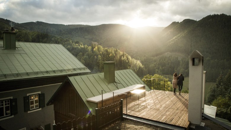 Two people stand on a terrace with a view of wooded mountains at sunset.