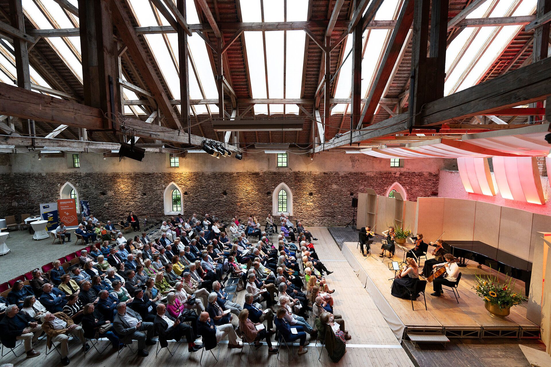 Konzert von „Harriet & Friends“ in der Alten Reitschule der Burg Feistritz. Ein Streichensemble spielt auf der Bühne vor einem großen Publikum in der historischen Halle mit offenem Dachstuhl und Natursteinmauern.