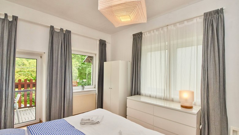 Bright bedroom with double bed, blue striped bedding, white furniture and gray curtains. Window overlooking a balcony.