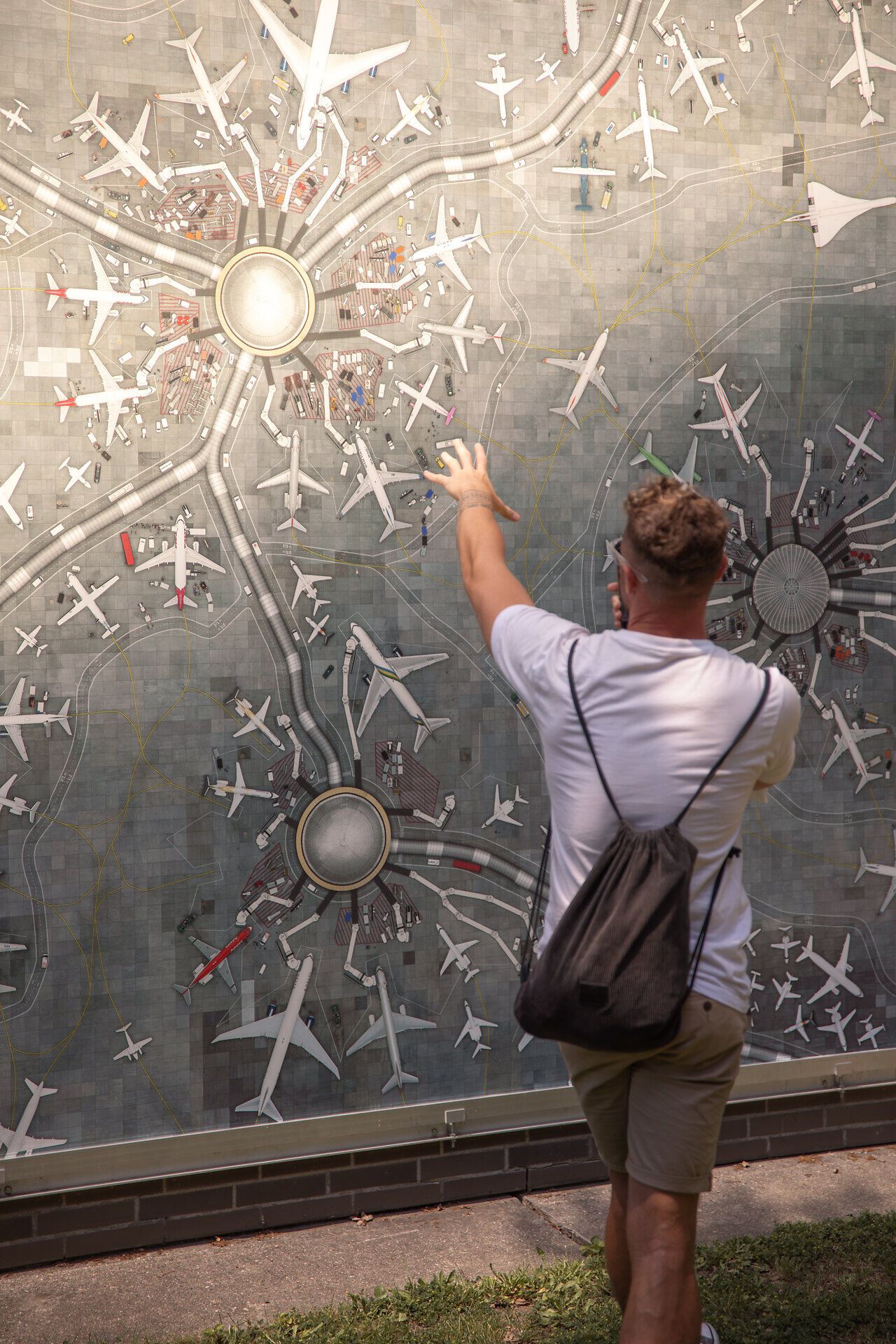 Ein begeisterter Besucher steht vor einer beeindruckenden Wandkarte, die die Flugzeuge und die Weite des Himmels darstellt. Mit einer Hand ausgestreckt, scheint er die verschiedenen Routen und Ziele zu erkunden, während die Sonne sanft auf die Szenerie scheint. Diese lebendige Darstellung weckt die Vorfreude auf neue Abenteuer und Entdeckungen.