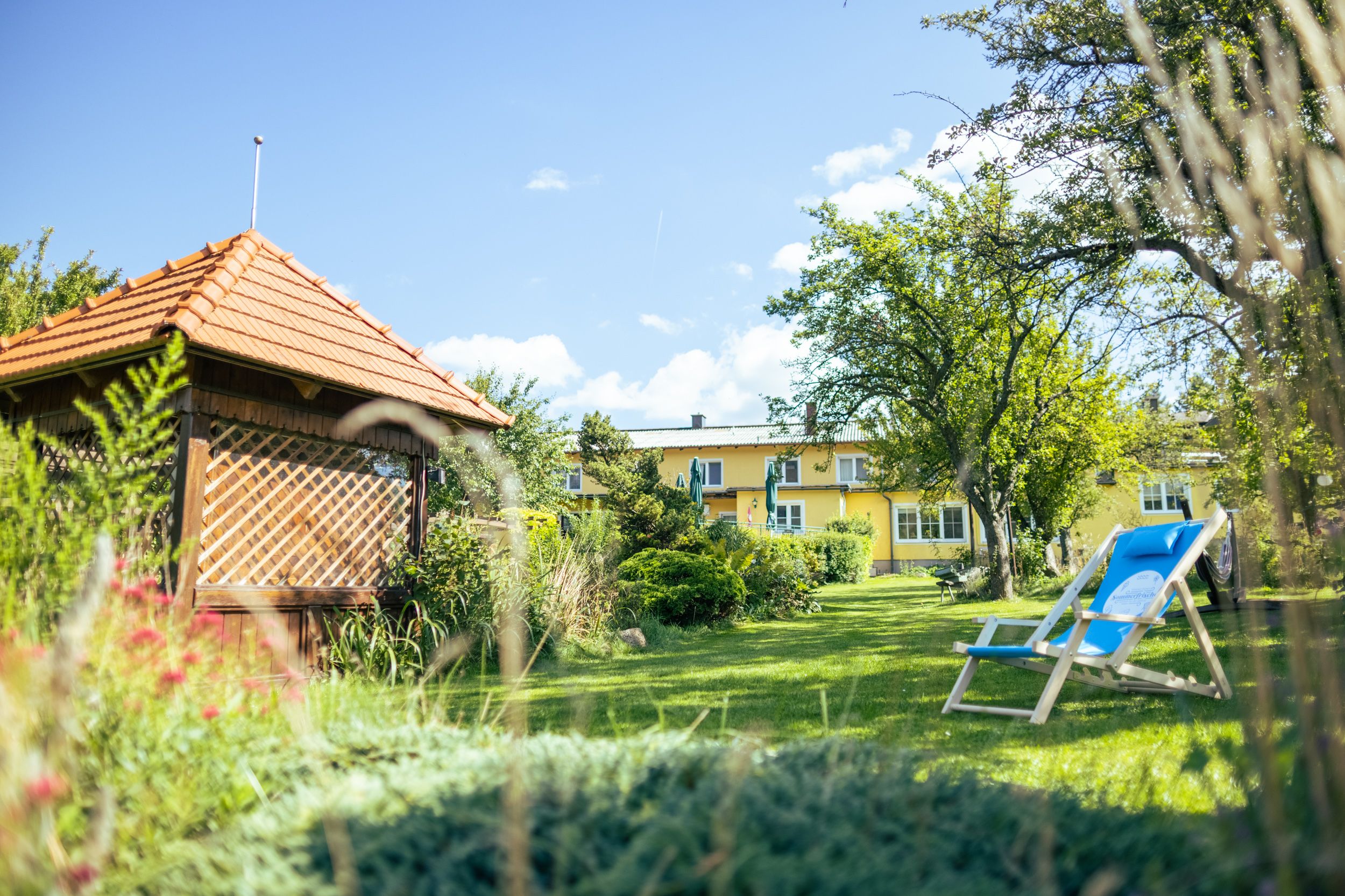 Ein grüner Garten mit einem Pavillon, Liegestuhl und einem gelben Gebäude im Hintergrund.