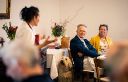 Eine Person steht in einer Gaststube und spricht mit zwei am Tisch sitzenden Personen. Ein Hund sitzt daneben und schaut die stehende Person an.