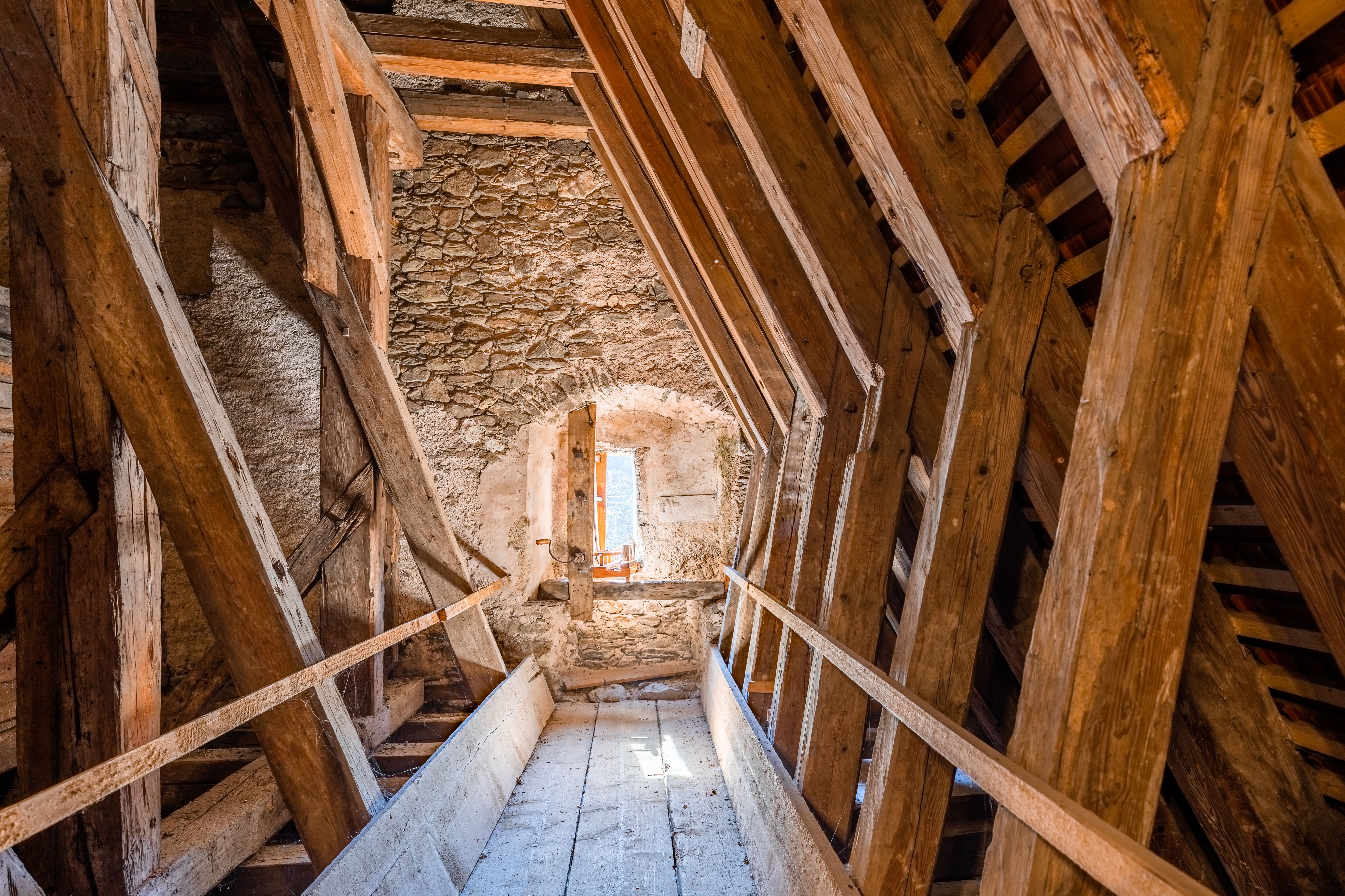 Dachgeschoss der Wehrkirche Edlitz mit vielen Holzbalken und Steinmauer, durch eine Fensteröffnung in der Mauer kommt Licht in den Raum.