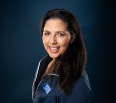 Portraitfoto Wiener Alpen Mitarbeiterin, die eine Jacke mit Wiener Alpen Logo trägt und vor einem dunklem Hintergrund steht.