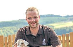 Ein Mann sitzt auf einer Holzbank, hält eine Ziege und lächelt in die Kamera.