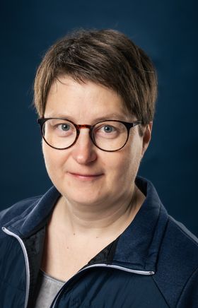 Portraitfoto Wiener Alpen Mitarbeiterin, die eine Jacke mit Wiener Alpen Logo trägt und vor einem dunklem Hintergrund steht.
