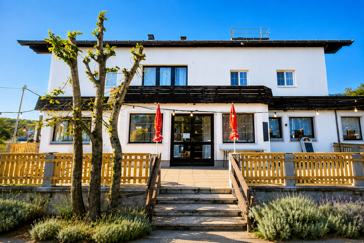 Außenansicht eines zweistöckigen Restaurants mit weißen Wänden und roten Sonnenschirmen vor dem Eingang.