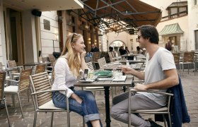 Zwei Personen sitzen an einem Tisch in einem Straßencafé und unterhalten sich.