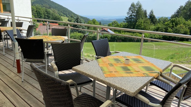 Terrasse mit Tischen und Stühlen, Blick auf grüne Hügel und Bäume im Hintergrund.