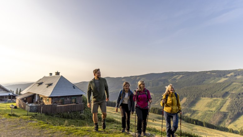 Four people hiking on a mountain path next to a hut at sunset.