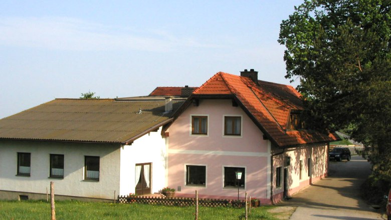 Ein traditionelles Gasthaus mit rosa und wei&szlig;en W&auml;nden und einem roten Ziegeldach, umgeben von gr&uuml;ner Landschaft.