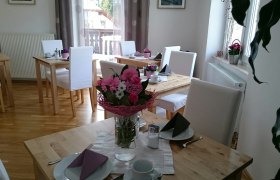 Ein heller Fr&uuml;hst&uuml;cksraum mit Holztischen, wei&szlig;en St&uuml;hlen und Blumenarrangements auf den Tischen.