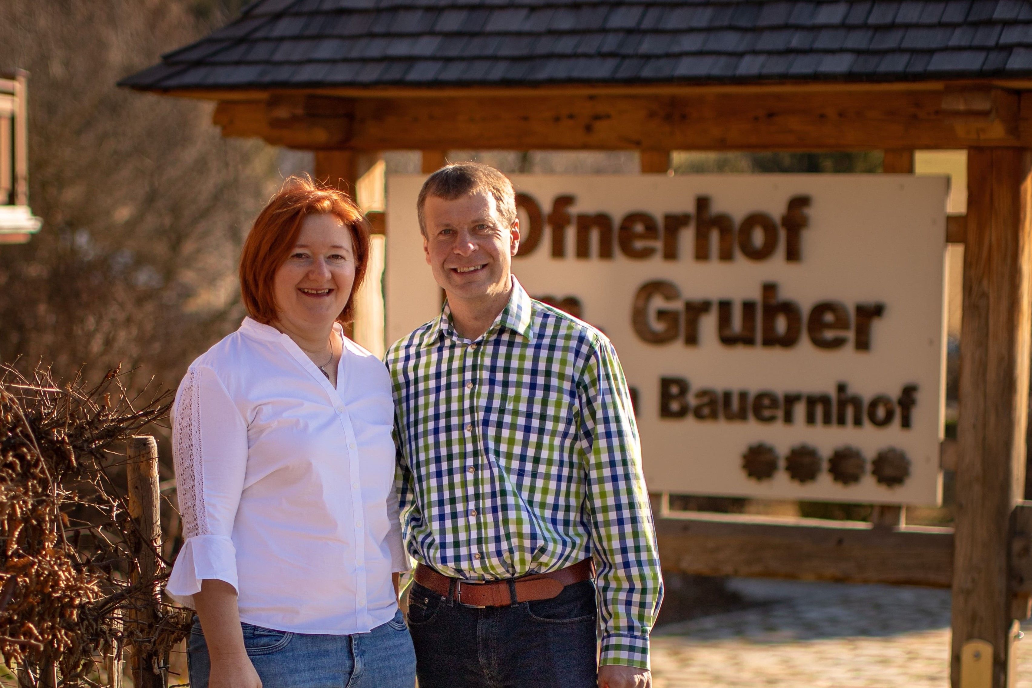 Ein Mann und eine Frau stehen lächelnd vor einem Schild mit der Aufschrift 'Ofnerhof Gruber Bauernhof'.