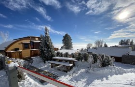 Winterliche Glamping-H&uuml;tte mit Schnee bedeckt, umgeben von B&auml;umen und blauem Himmel.