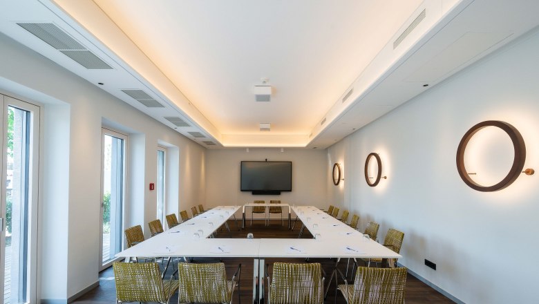 Moderner Seminarraum mit U-f&ouml;rmiger Tischanordnung, St&uuml;hlen, einem gro&szlig;en Bildschirm und dekorativen Wandlampen.