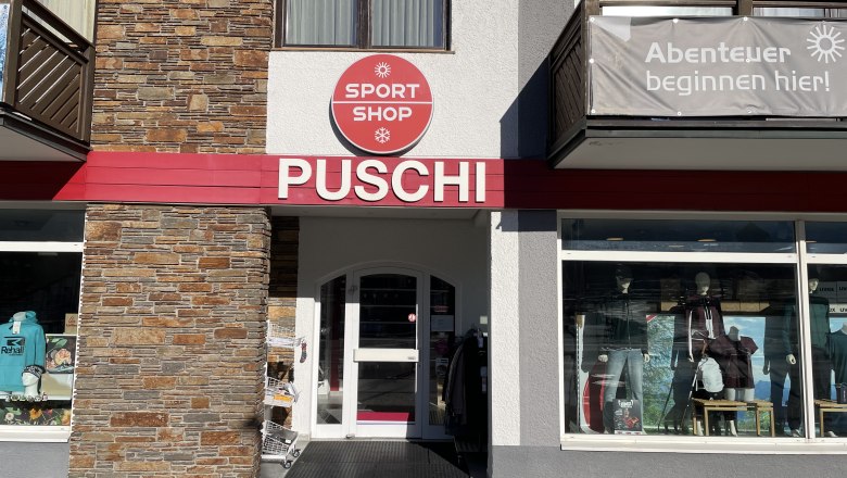 Eingangsbereich eines Sportgeschäfts mit der Aufschrift 'PUSCHI' und einem Schild 'SPORT SHOP'. Schaufenster mit Sportbekleidung und Ausrüstung.