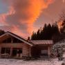 Ein Holzchalet im Winter mit schneebedecktem Boden und einem dramatischen, orangefarbenen Himmel im Hintergrund.
