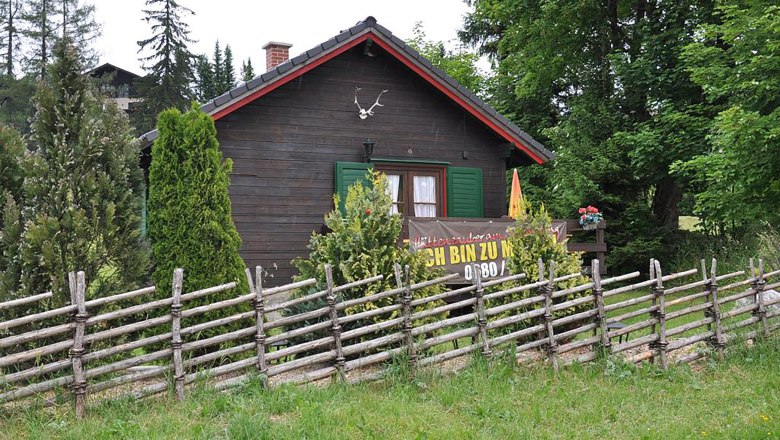 A rustic wooden hut with green shutters and a wooden fence in the foreground, surrounded by trees and shrubs.