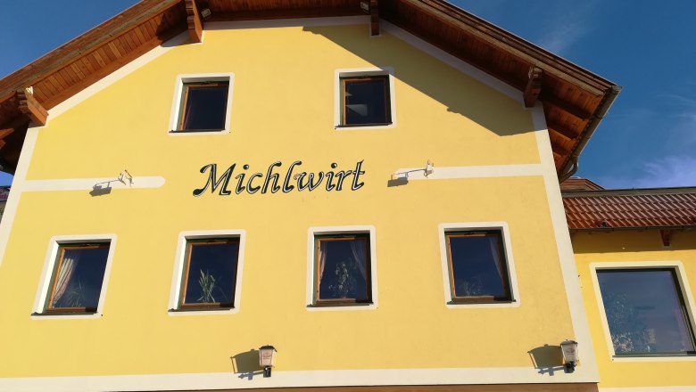 Gelbes Gebäude mit der Aufschrift 'Michlwirt' und mehreren Fenstern, unter blauem Himmel.