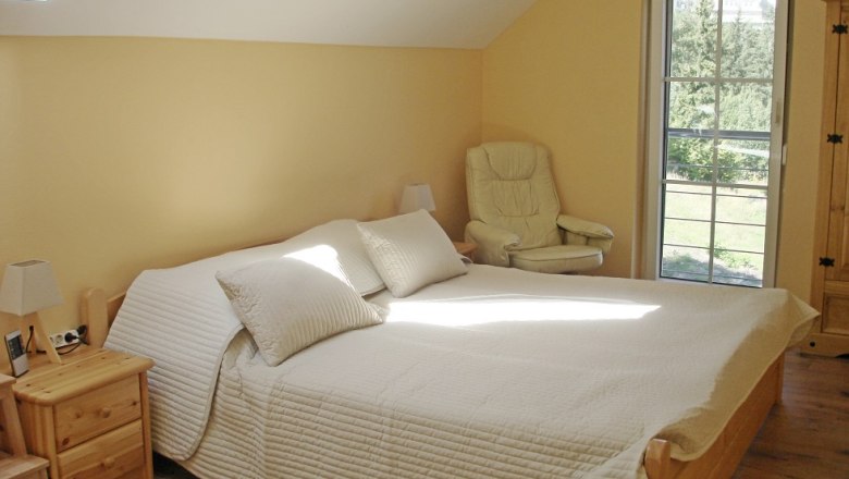 Helles Schlafzimmer mit Doppelbett, Sessel und Fenster mit Blick ins Grüne.