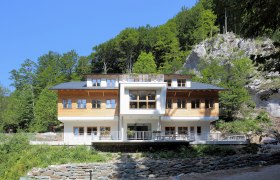 Ein modernes Gebäude mit Holzfassade vor einem bewaldeten Hang.