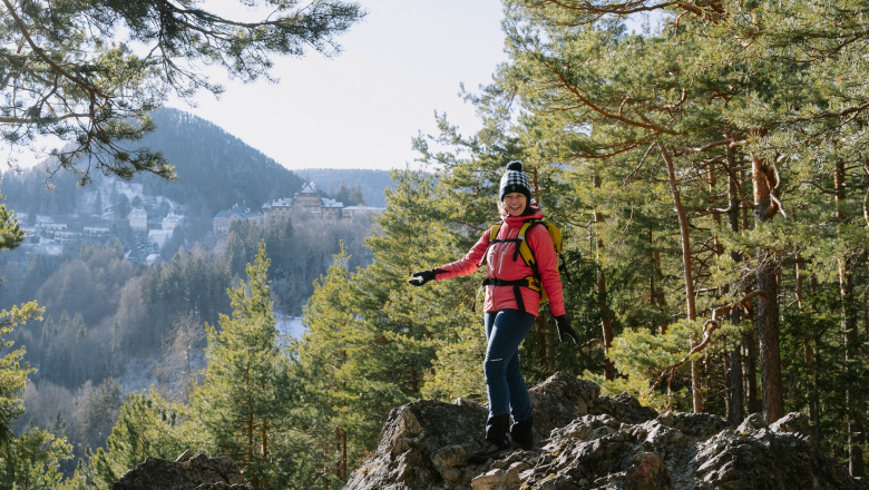 Person in pinker Jacke wandert auf einem felsigen Pfad im Wald mit Bergen im Hintergrund.