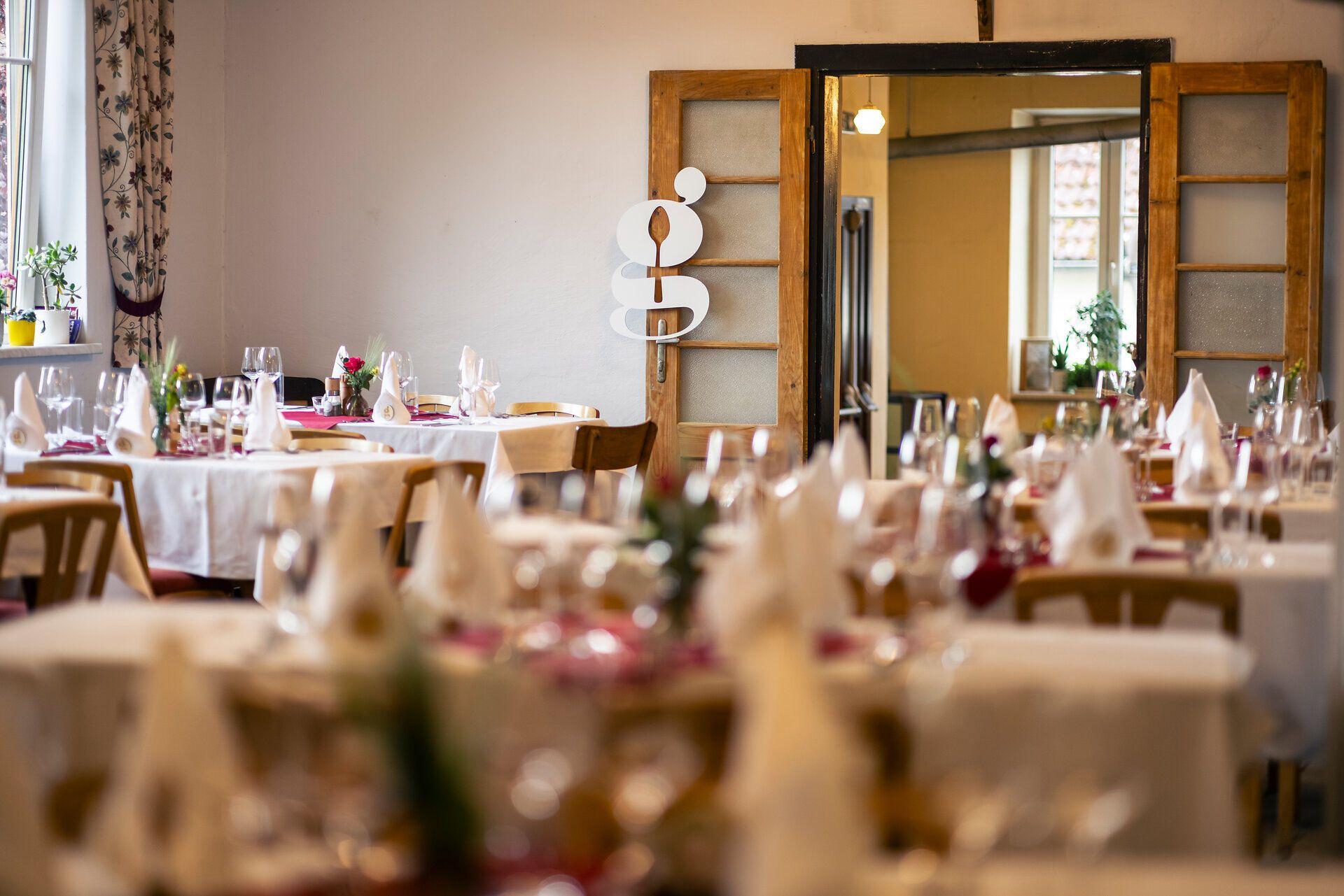 Gaststube im Gasthaus Stocker mit vielen gedeckten Tischen zur Veranstaltung Gedanken-Gänge. An der doppelflügigen Tür hängt ein kleines G, das Logo der Veranstaltung.