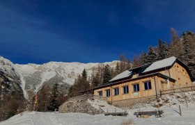 A wooden house in the snow against a mountain backdrop under a clear blue sky.