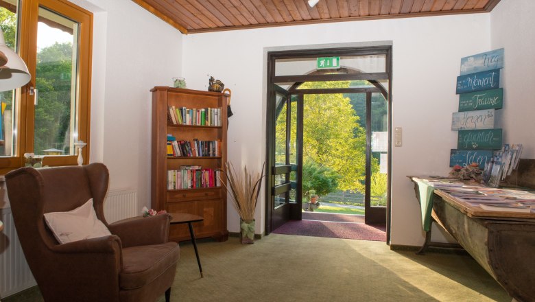 Eingangsbereich mit Sessel, B&uuml;cherregal und offenem T&uuml;rbereich mit Blick ins Gr&uuml;ne.