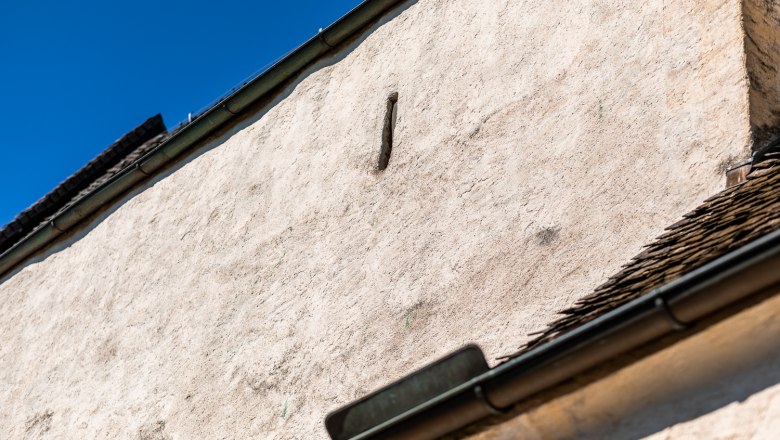 Nahaufnahme einer weißen Mauer mit Schießscharte Dachrinne und blauen Himmel im Hintergrund.