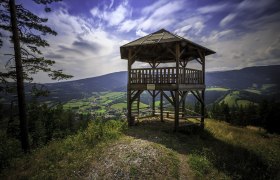 Holzaussichtsturm mit Blick auf grüne Täler und Berge unter blauem Himmel.