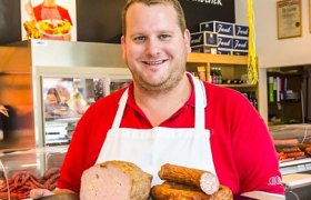 A butcher in a red shirt and white apron holds various sausage products in a butcher's shop.